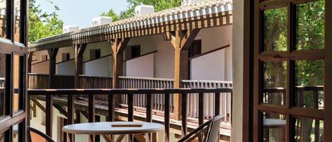 Un balcón con mesa y sillas, vigas de madera y vista a árboles y edificios.