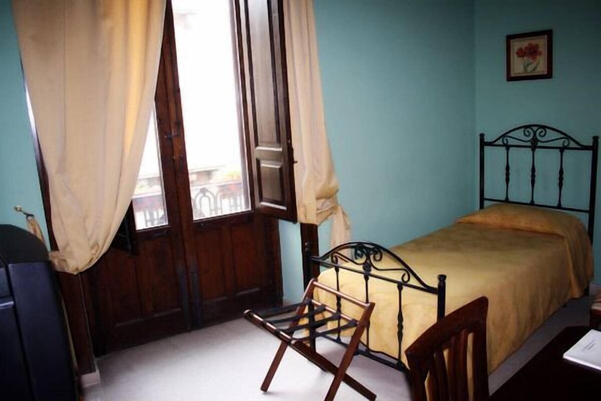 Ein Schlafzimmer mit Bett, Sessel, Schreibtisch, einem Fenster mit Vorhängen und einem Bild an der Wand.