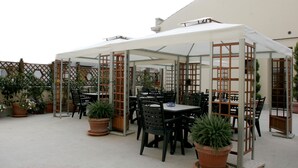 Un espace extérieur aménagé en terrasse, avec plusieurs tables et chaises, protégé par un auvent blanc et entouré de plantes en pot et d’une clôture en treillis de bois.