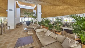 Un área de sala de estar interior con techo alto, ventanas amplias y varios conjuntos de asientos, incluyendo sofás y mesas.