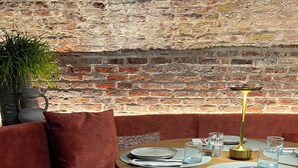 Un rincón acogedor con un banco tapizado rojo, una mesa redonda de madera dispuesta con platos, vasos y un candelabro.