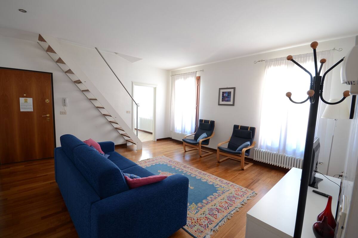 Un salón amplio con un sofá azul, una escalera de madera y vistas al exterior a través de ventanas grandes.