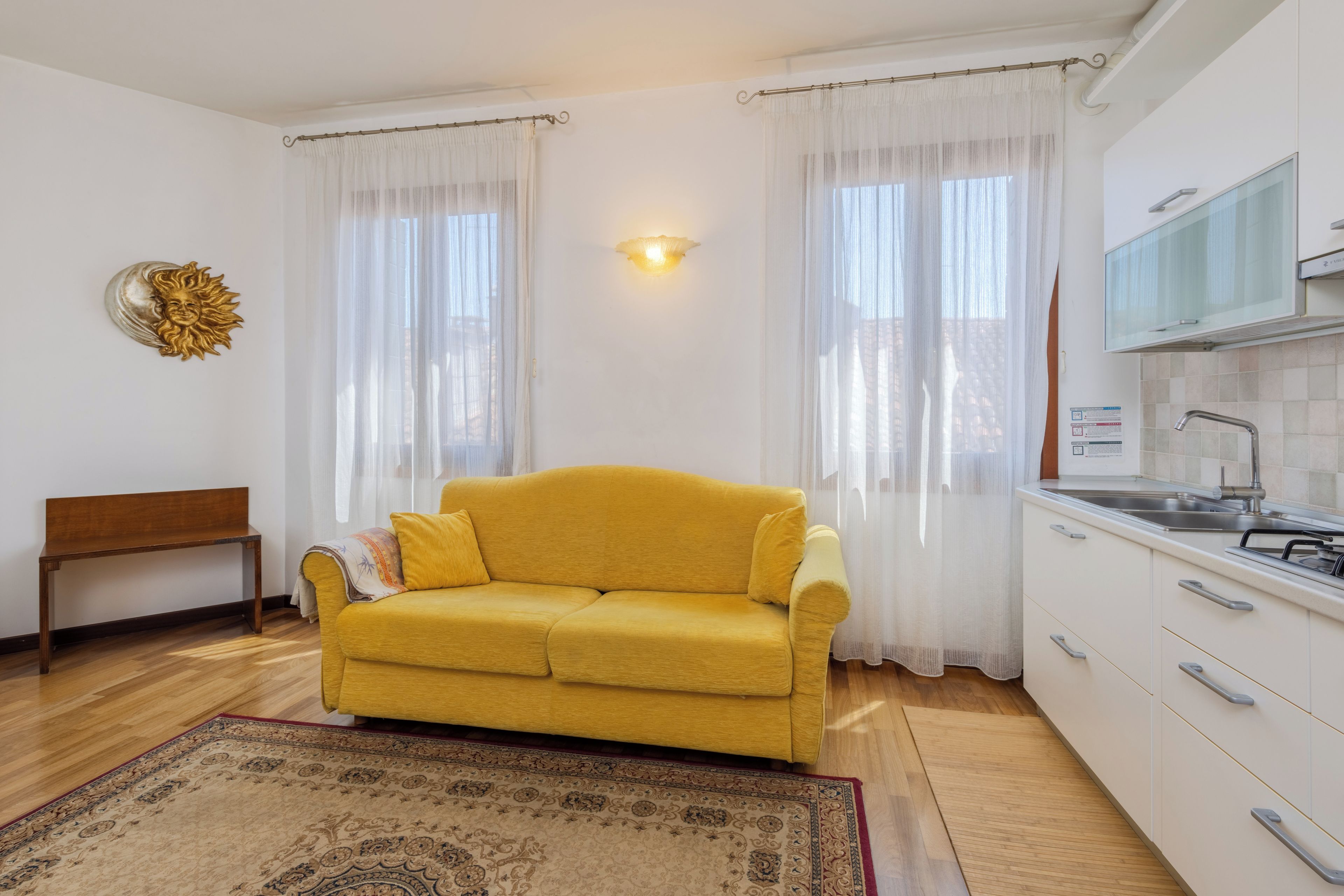 Un divano giallo in una stanza con angolo cottura, una panca e un tappeto decorato.