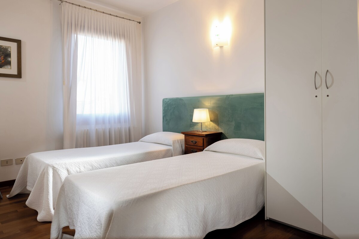 A hotel room with two single beds, a nightstand with a lamp, a wardrobe, and a window with curtains.