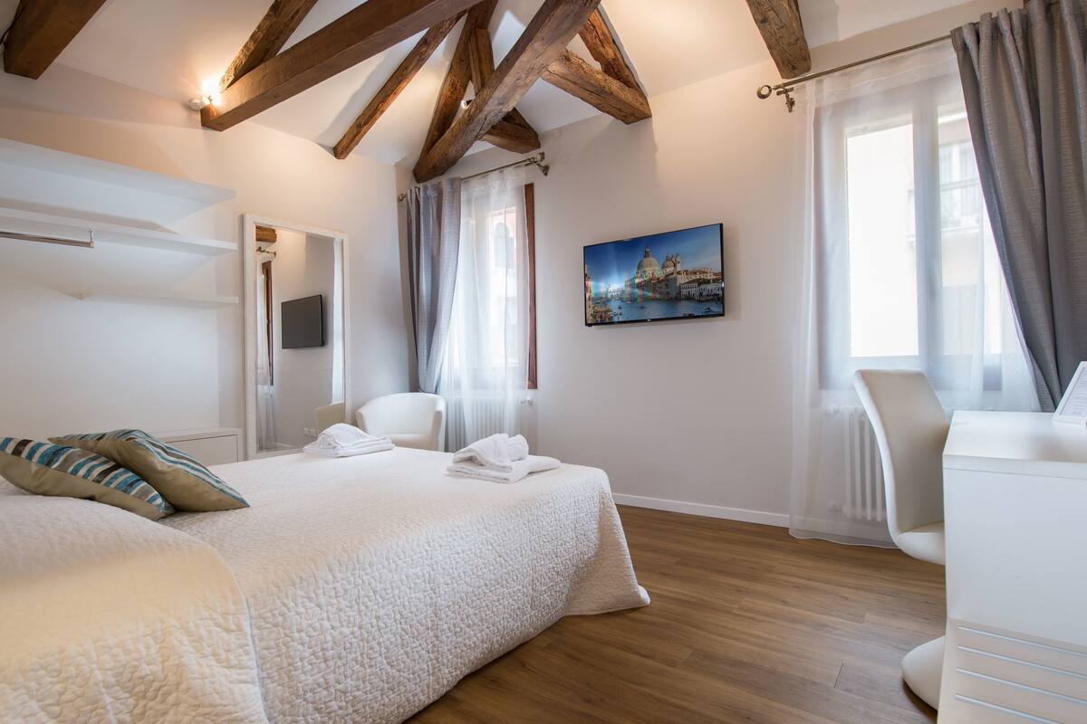 Un dormitorio con una cama grande, un escritorio y una silla. Hay un cuadro en la pared y una ventana con cortinas.
