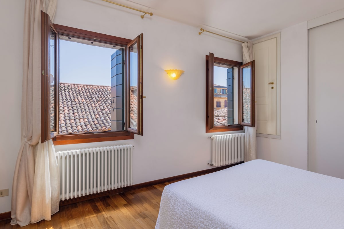 A room with a bed, two windows with curtains, a wall-mounted light, and a radiator.