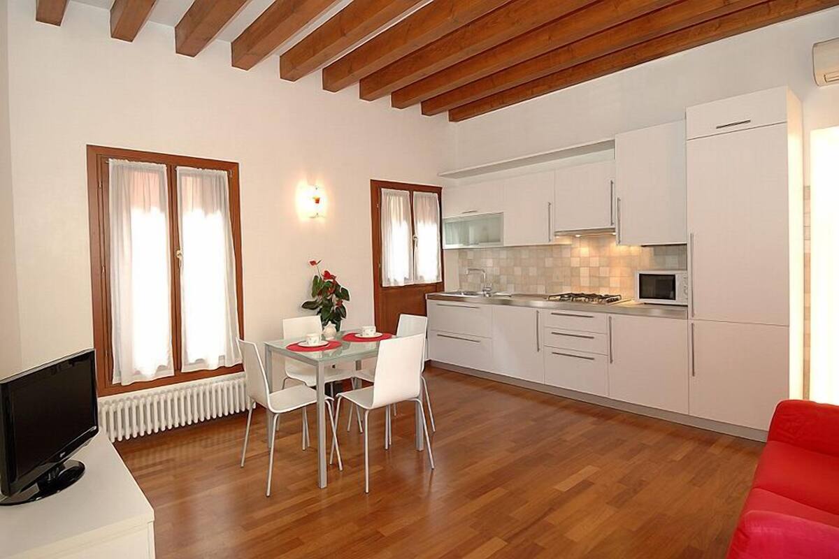 Una cocina moderna con armarios blancos, un microondas y una estufa a gas. Hay un comedor con una mesa y sillas, y un sofá rojo en la esquina.