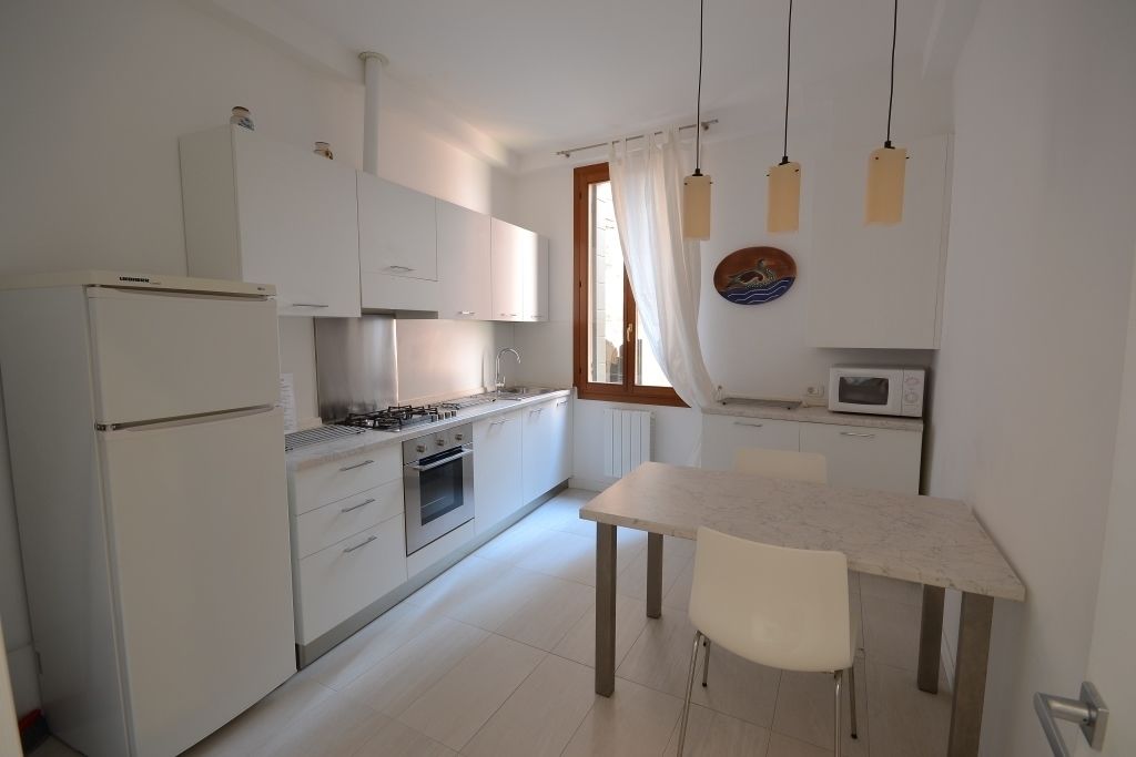 Une cuisine moderne avec des meubles blancs, une table à manger avec des chaises, un four encastré, un four à micro-ondes et des luminaires suspendus.