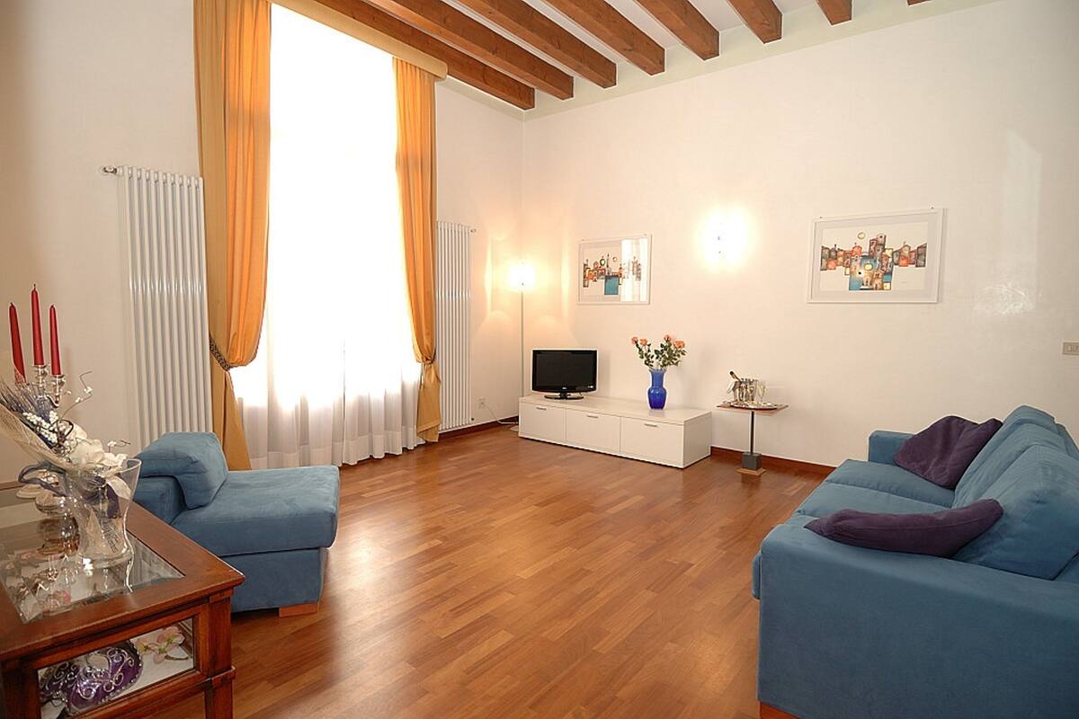 Una sala de estar con dos sofás azules, un televisor sobre un mueble blanco, una mesa de centro de vidrio y un florero con flores.