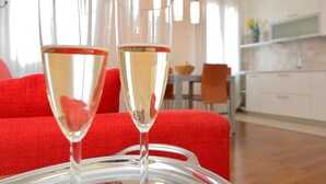 Dos copas de vino blanco sobre una mesa de vidrio, con un corcho en el centro, en un salón moderno con un sofá rojo y una cocina al fondo.