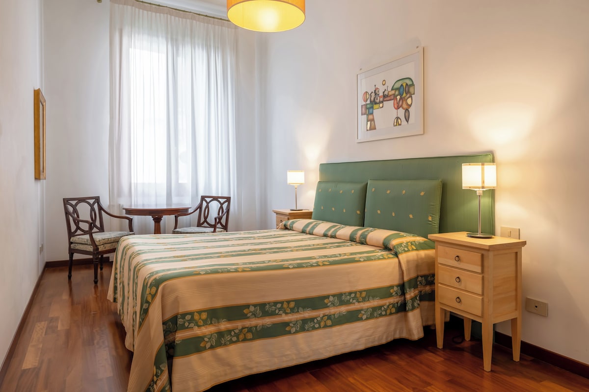 A hotel room with a bed, two bedside tables, a chair, a small table, and a window with curtains.