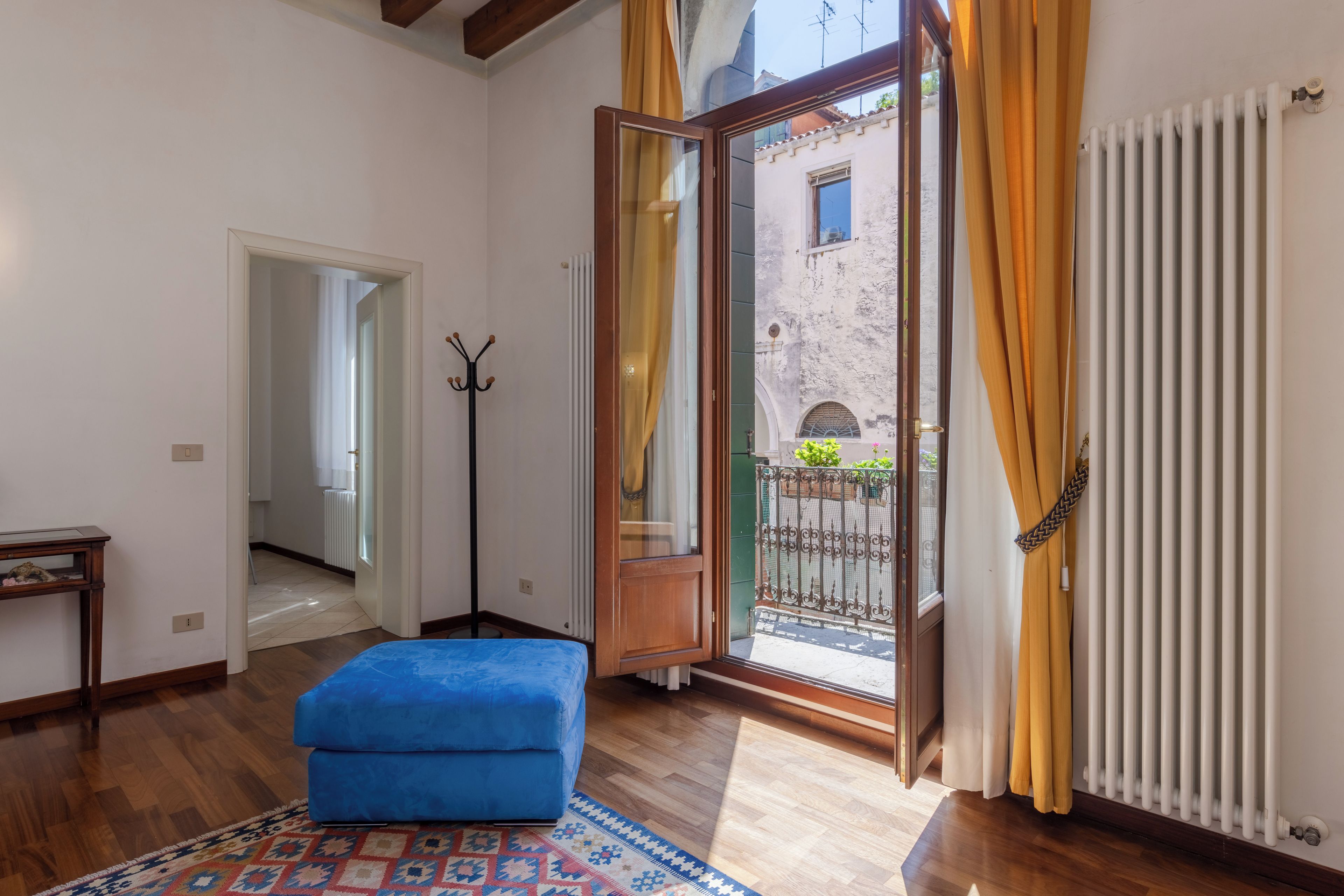 Una stanza con un pouf blu, un termosifone, un appendiabiti e vista su un edificio attraverso una porta vetrata.