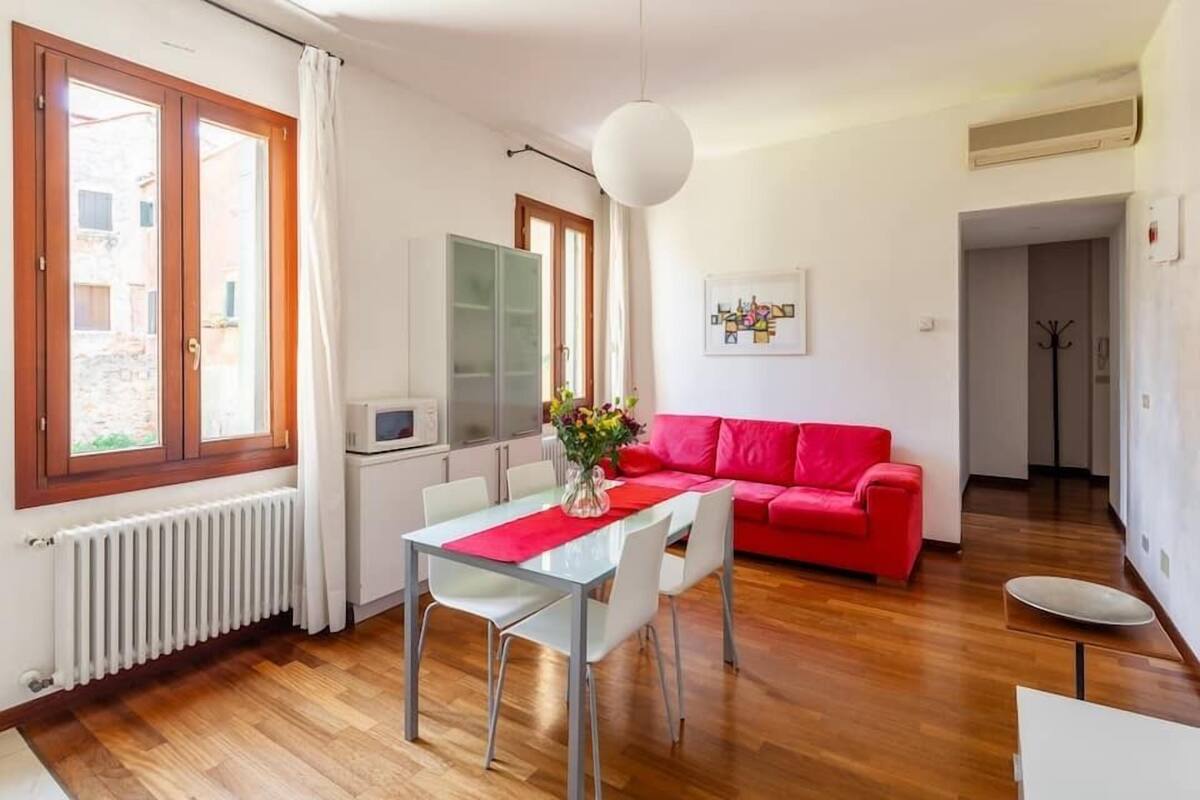 Una sala de estar moderna con un sofá rojo, una mesa de comedor de vidrio y una zona de cocina con microondas.