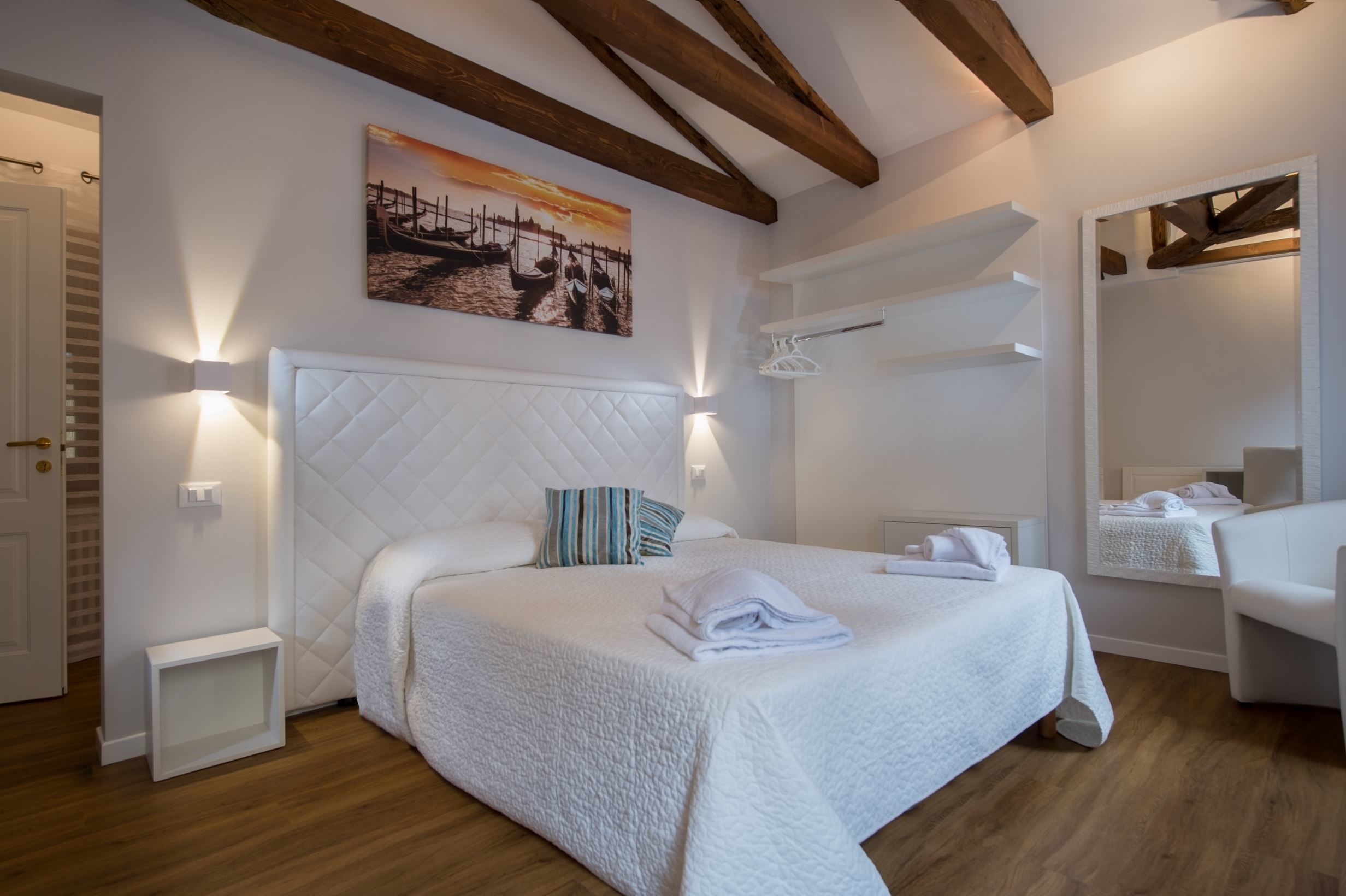 Une chambre d’hôtel avec un grand lit, un plafond en bois, un tableau représentant des bateaux et un miroir au mur.
