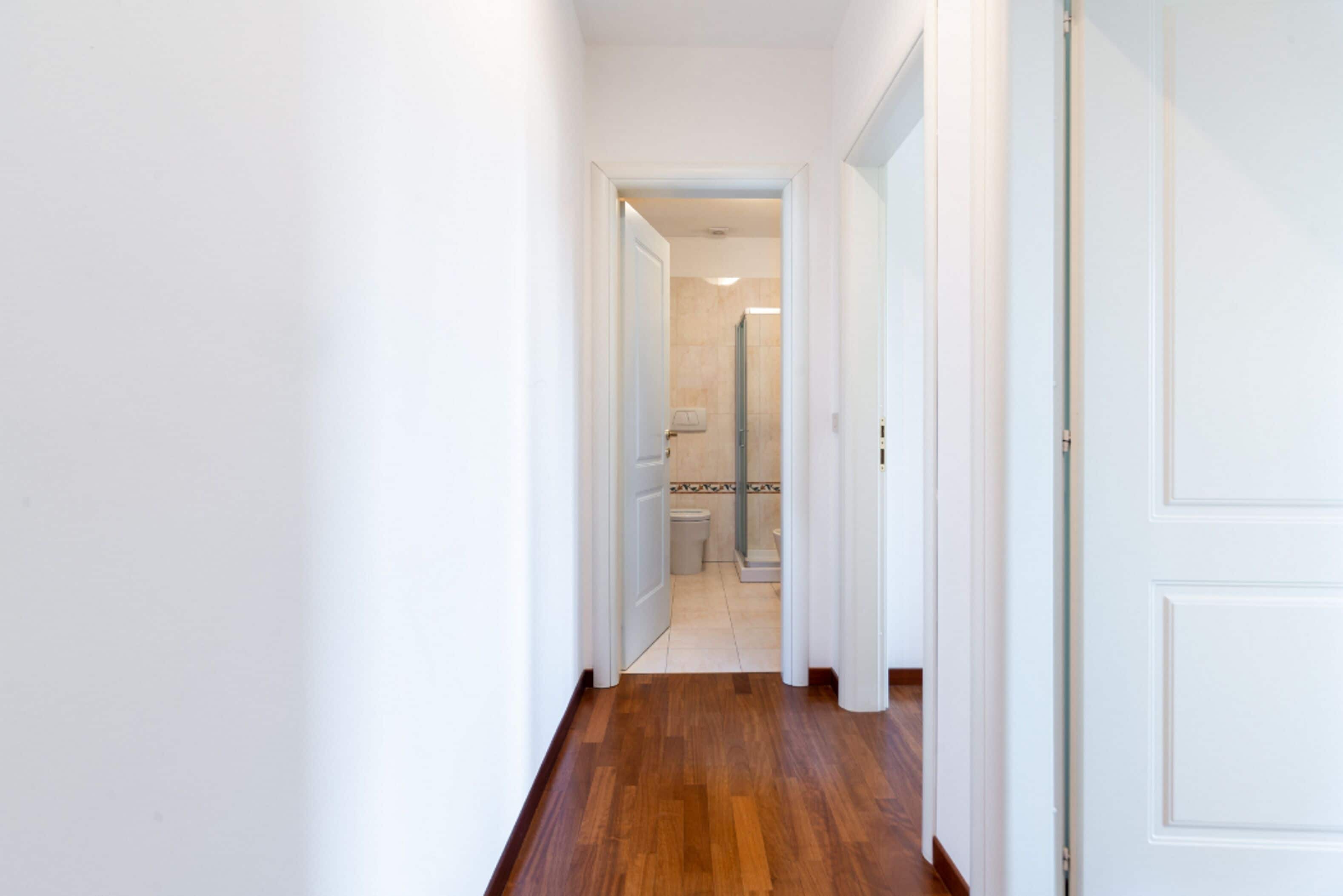 Un couloir étroit, doté d’un plancher en bois et de murs blancs, qui mène à une salle de bain avec douche et toilettes.