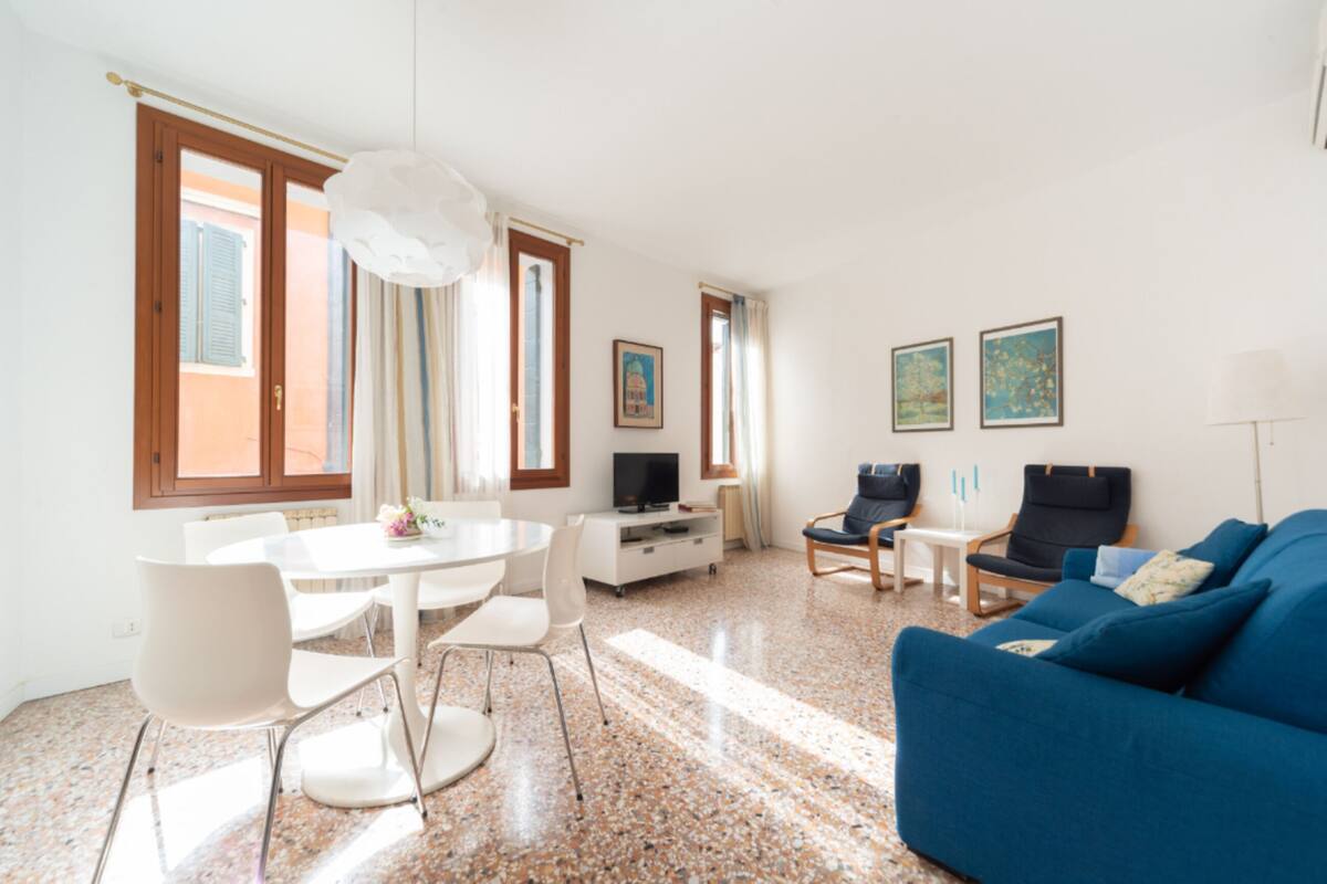 Un salón moderno con un sofá azul, una mesa de comedor blanca y sillas. Hay un televisor sobre un mueble blanco, y obras de arte en las paredes.