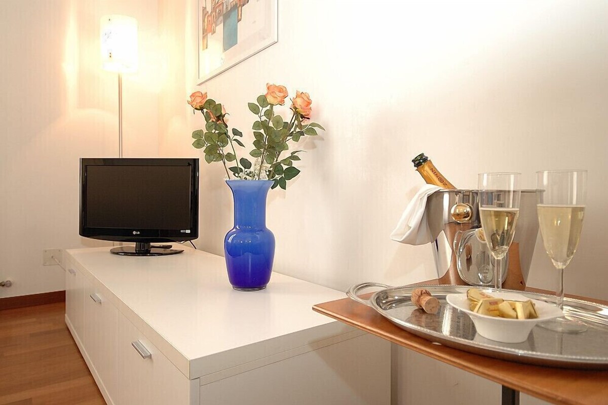 A hotel room with a flat-screen TV, a blue vase with flowers, a tray with glasses of wine, a bottle of champagne, and a bowl of fruit.
