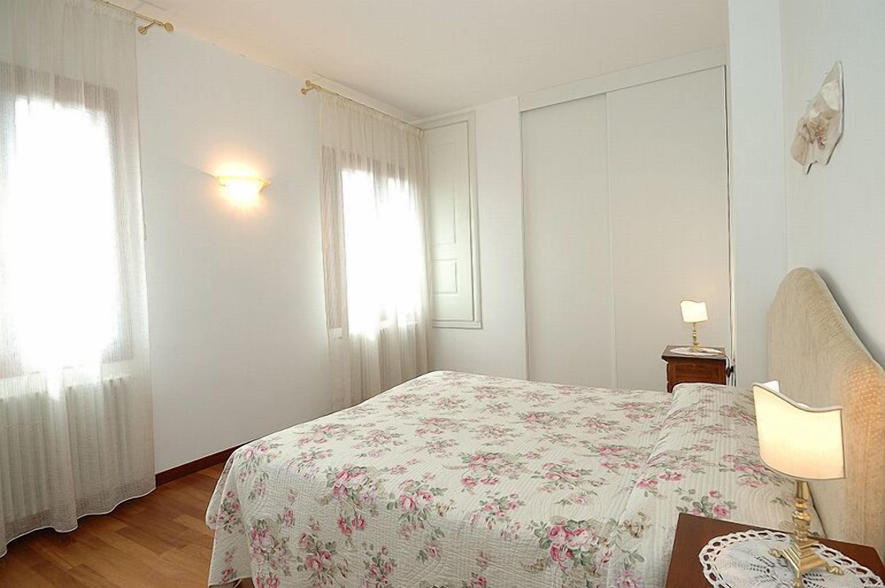 Une chambre à coucher avec une couvre-lit à motifs floraux, une table de chevet avec une lampe et une fenêtre ornée de rideaux légers.