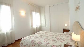 Une chambre à coucher avec une couvre-lit à motifs floraux, une table de chevet avec une lampe et une fenêtre ornée de rideaux légers.