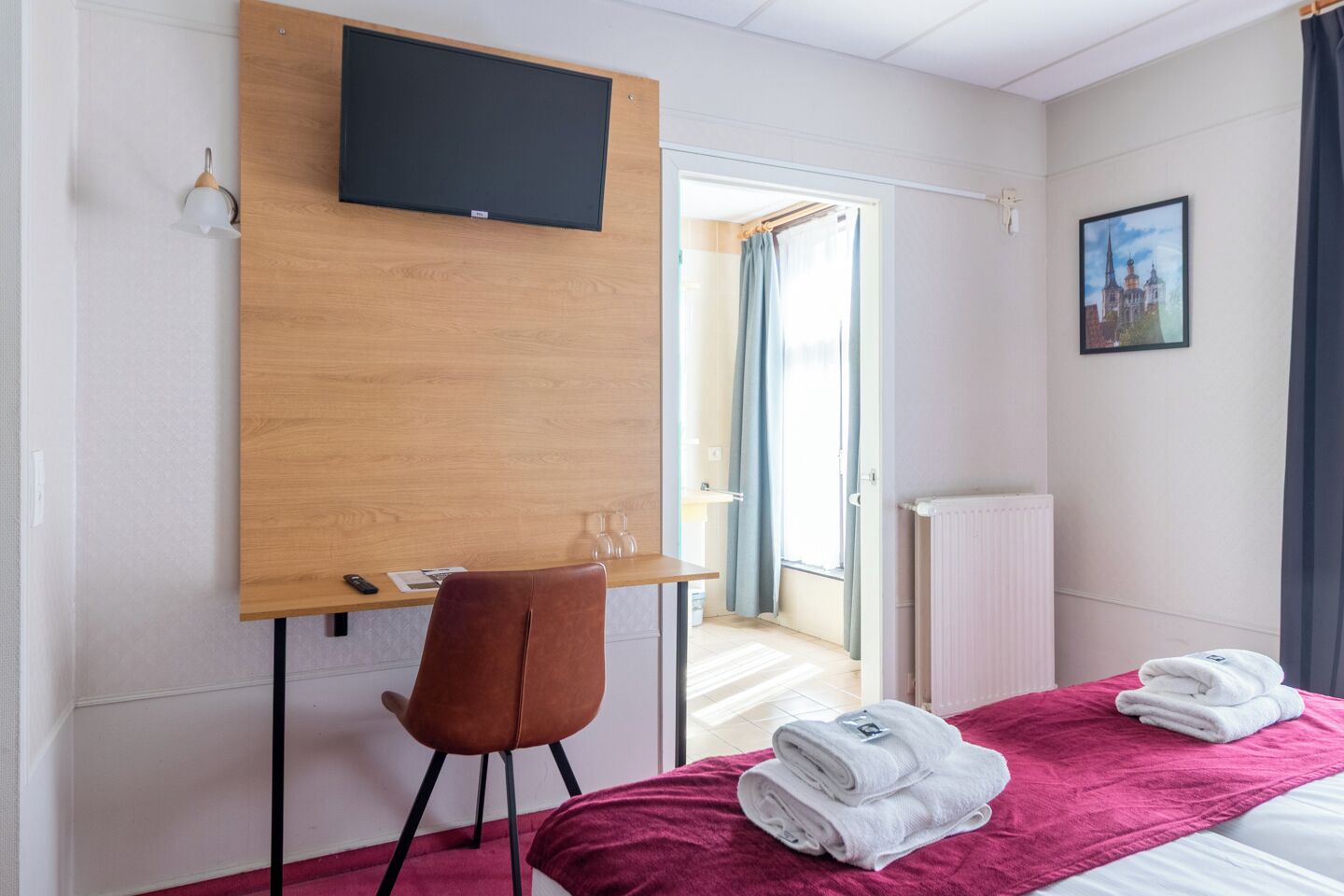 Quarto de hotel com cama, escrivaninha e cadeira, TV fixada na parede, radiador e janela com cortinas.