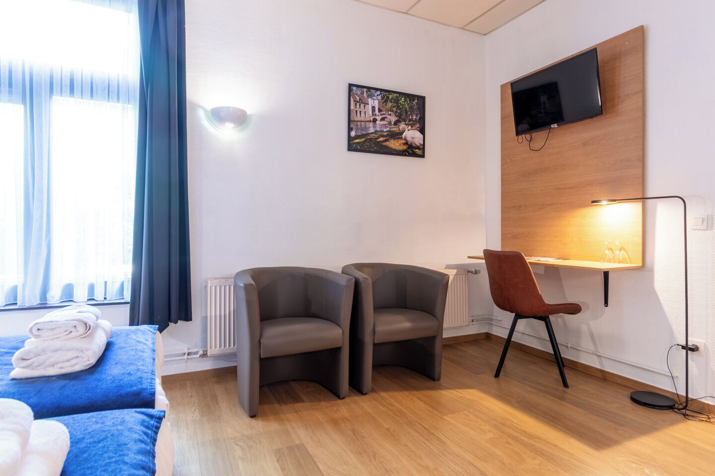Una habitación de hotel moderna con cama, dos sillones, un escritorio, televisor montado en la pared y una ventana con cortinas.