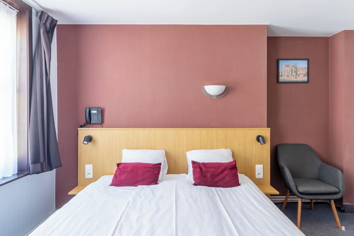 A hotel room with a wooden headboard, two white beds with red pillows, a grey armchair, a wall-mounted phone, and a painting on the wall.