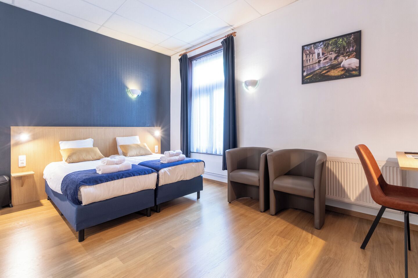 A modern hotel room with a large bed, two bedside tables, a desk with a chair, and a large window with curtains.
