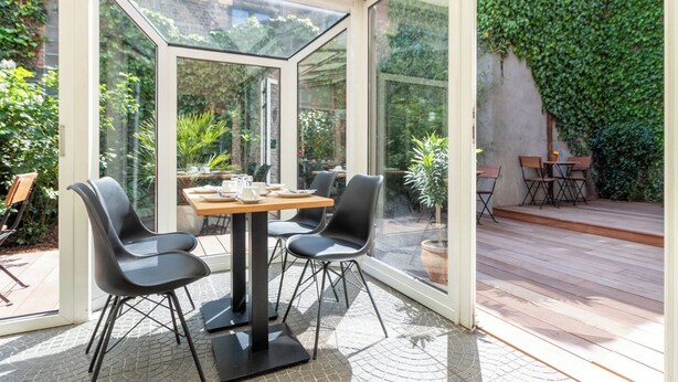 Une véranda avec une table et des chaises, entourée de murs de verre et offrant une vue sur la verdure.