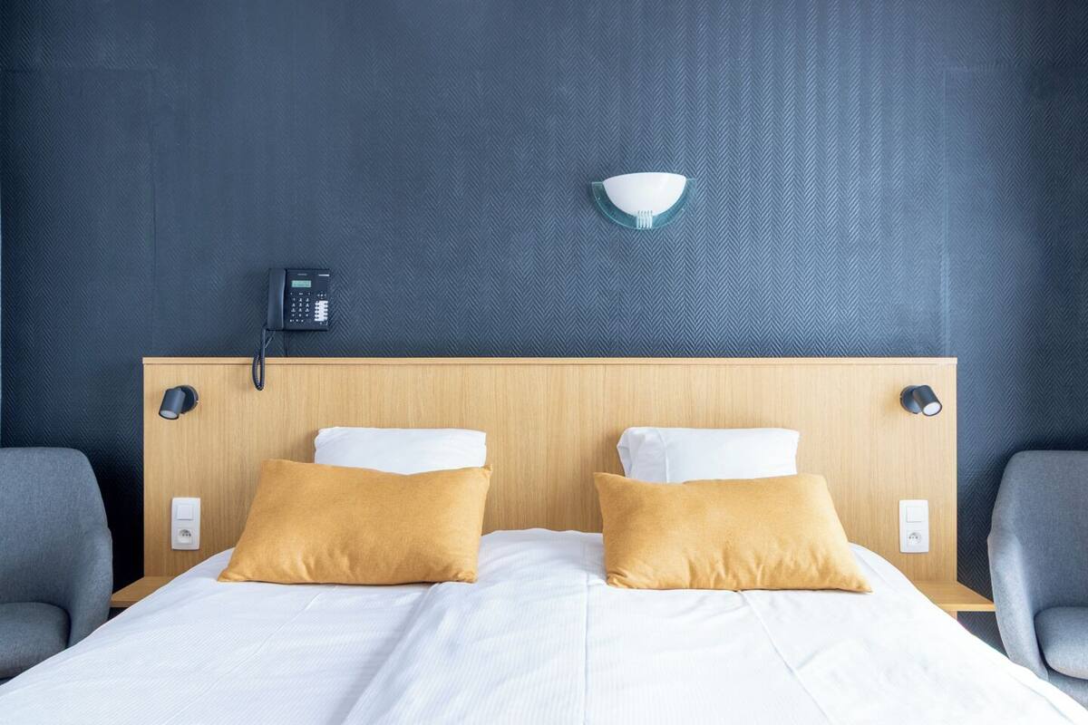 A hotel room with a wooden headboard, two white pillows, a light switch, a phone, and a wall-mounted light fixture.