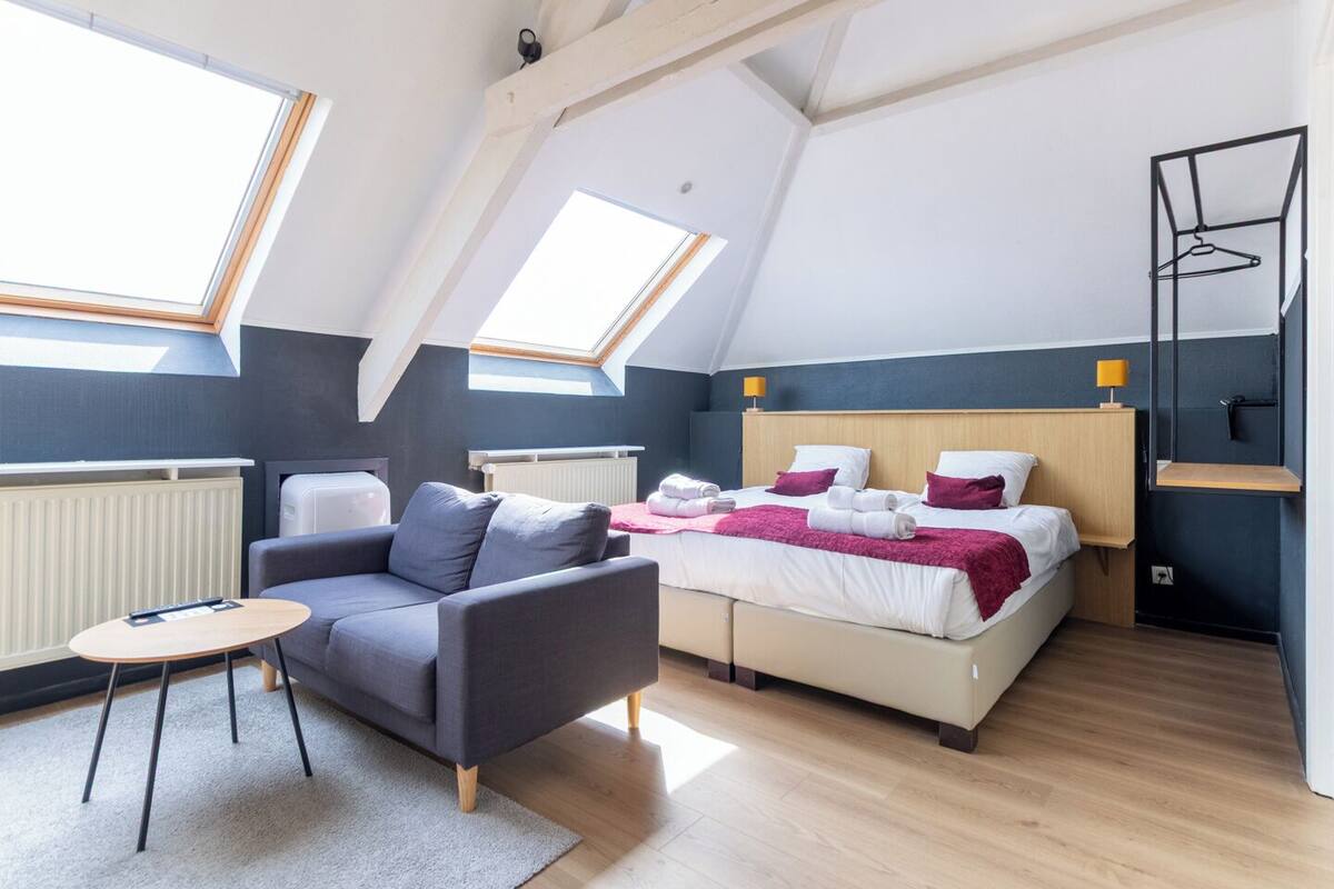 A modern loft bedroom with skylights, a bed with white and pink bedding, a grey sofa, and a small round table.