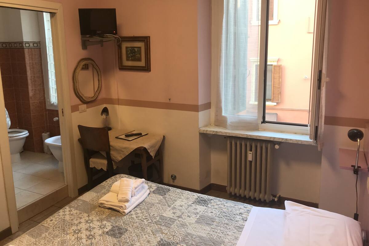 A hotel room with a bed, a small table, a chair, a window with a view, a radiator, and a bathroom visible through an open door.