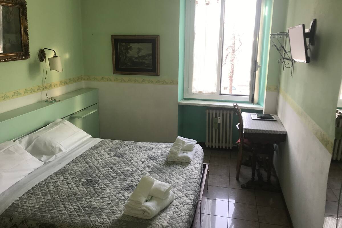 A room with a bed, a desk, a chair, a radiator, a window, and a wall-mounted TV.