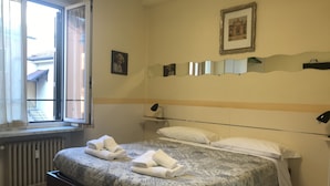 Habitació d'hotel amb un llit, una finestra, un radiador i una estanteta penjada a la paret amb una fotografia emmarcada.
