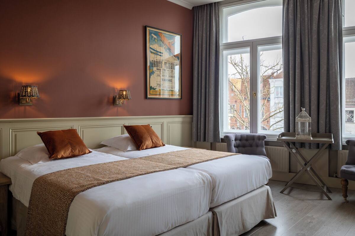 A hotel room with a large bed, two pillows, a nightstand, a table with a birdcage, and a window with curtains.