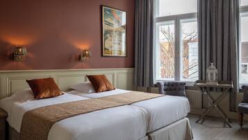 A hotel room with a large bed, two pillows, a nightstand, a table with a birdcage, and a window with curtains.