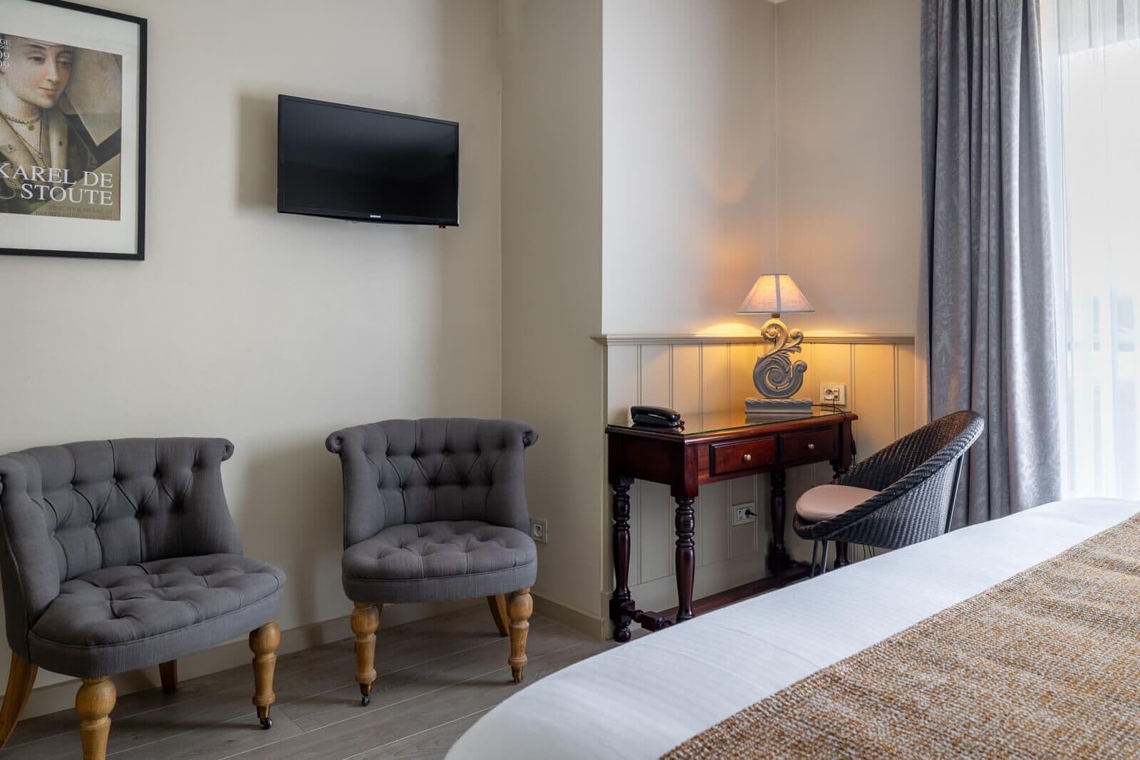 A hotel room with a bed, two armchairs, a desk with a lamp, a television, and a framed picture on the wall.