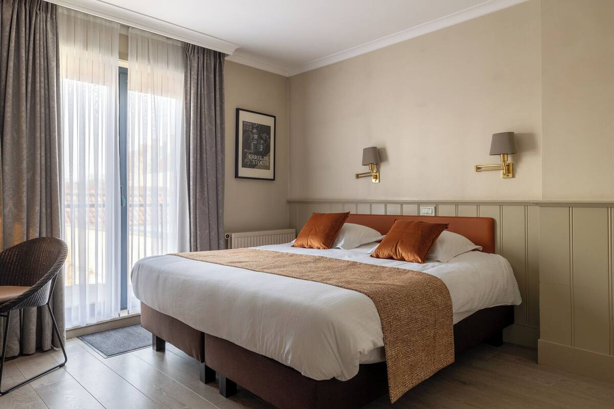 A hotel room with a large bed, two wall-mounted lamps, a framed picture, and a chair by the window.