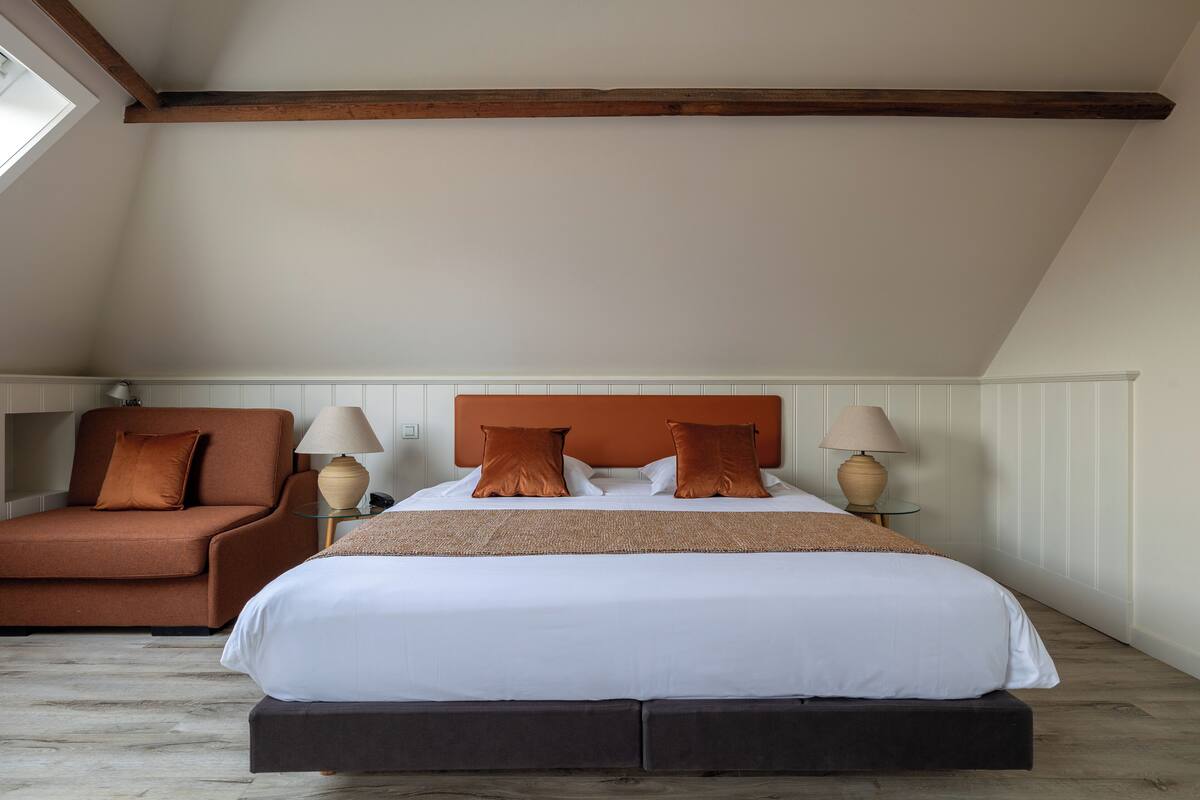 A bedroom with a large bed, a brown armchair, two bedside lamps, and a wooden ceiling beam.