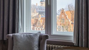 A room with a view of buildings through a large window, a chair with a cushion, and a small round table. - Ter Duinen (Bruges)