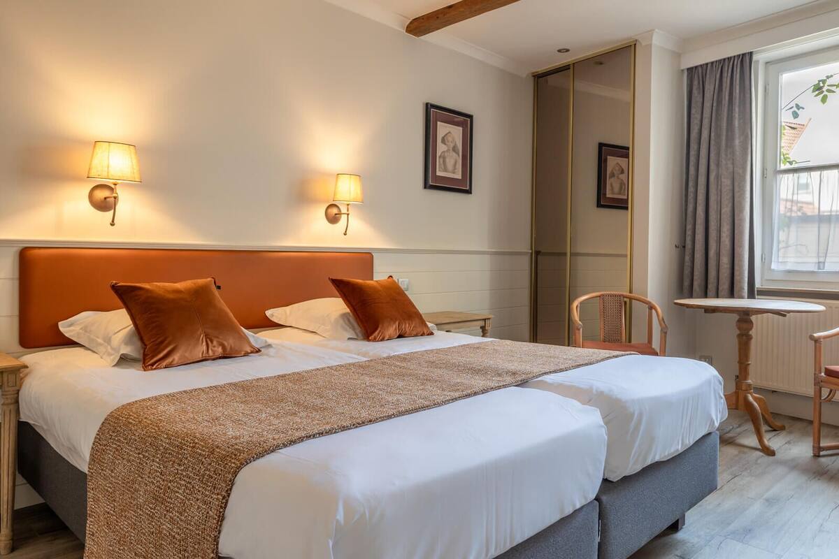 A hotel room with a double bed, two wall-mounted lamps, a small table, a chair, and a mirror.
