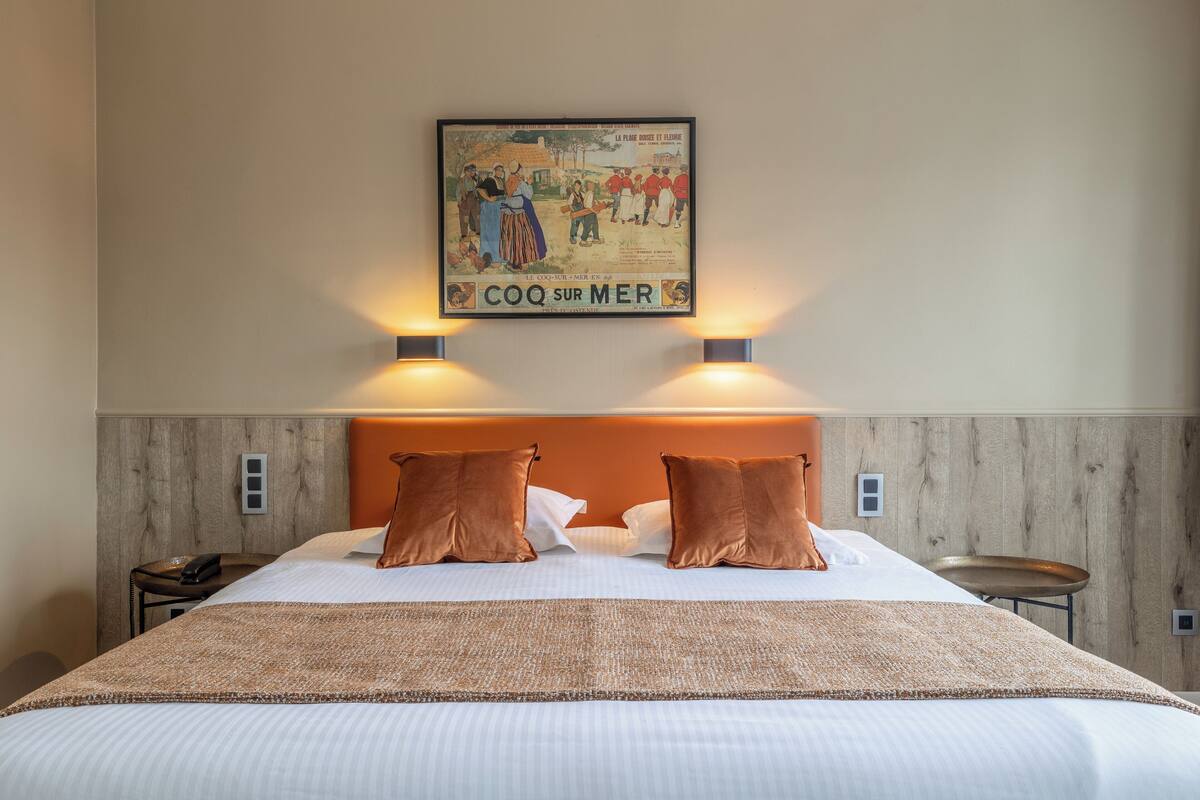 A hotel room with a large bed, two brown pillows, a headboard, a bedside table with a phone, and a framed picture on the wall.