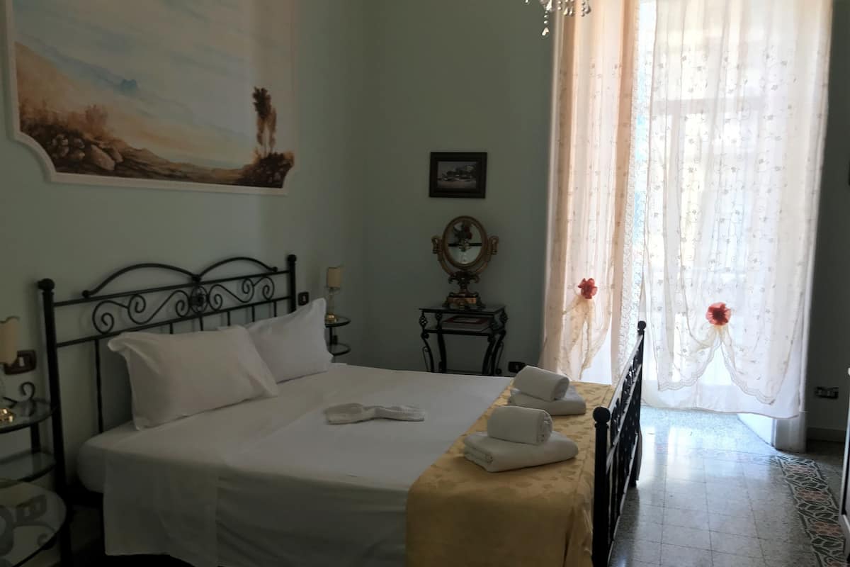A neatly made bed with white linens, a bedside table with a lamp, a small table with a mirror, and a window with sheer curtains.