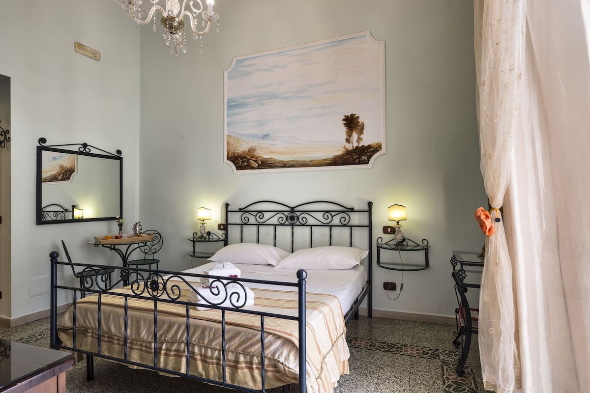 A bedroom with a metal bed frame, a bedside table with a lamp, a wall-mounted mirror, and a painting of a landscape.