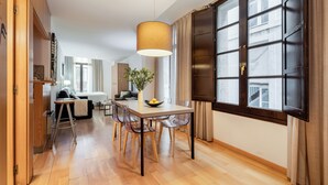 Et moderne spiseområde med et træbord, gennemsigtige stole, en hængende lampe og et stort vindue med gardiner.