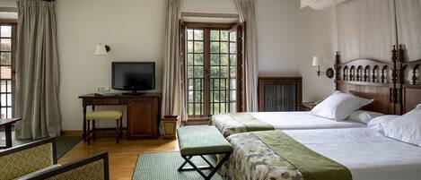 Habitación de hotel con dos camas, un escritorio con televisor, un banco y vista al exterior a través de una ventana.