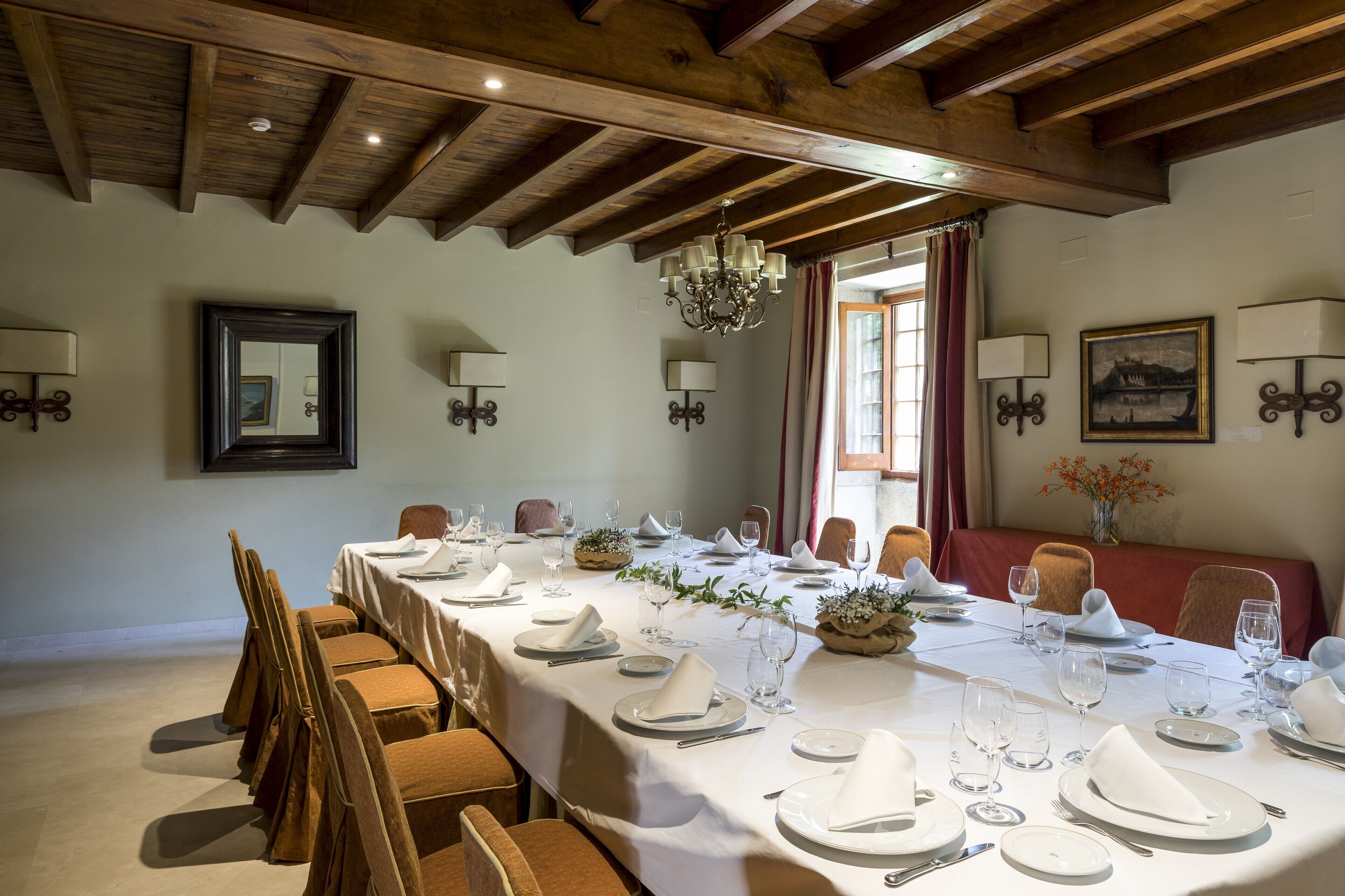 Un comedor con una mesa larga dispuesta para una comida, vigas de madera en el techo y lámparas fijadas a la pared.