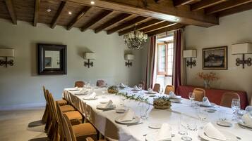 A dining room with a long table set for a meal, wooden beams on the ceiling, and wall-mounted lamps.