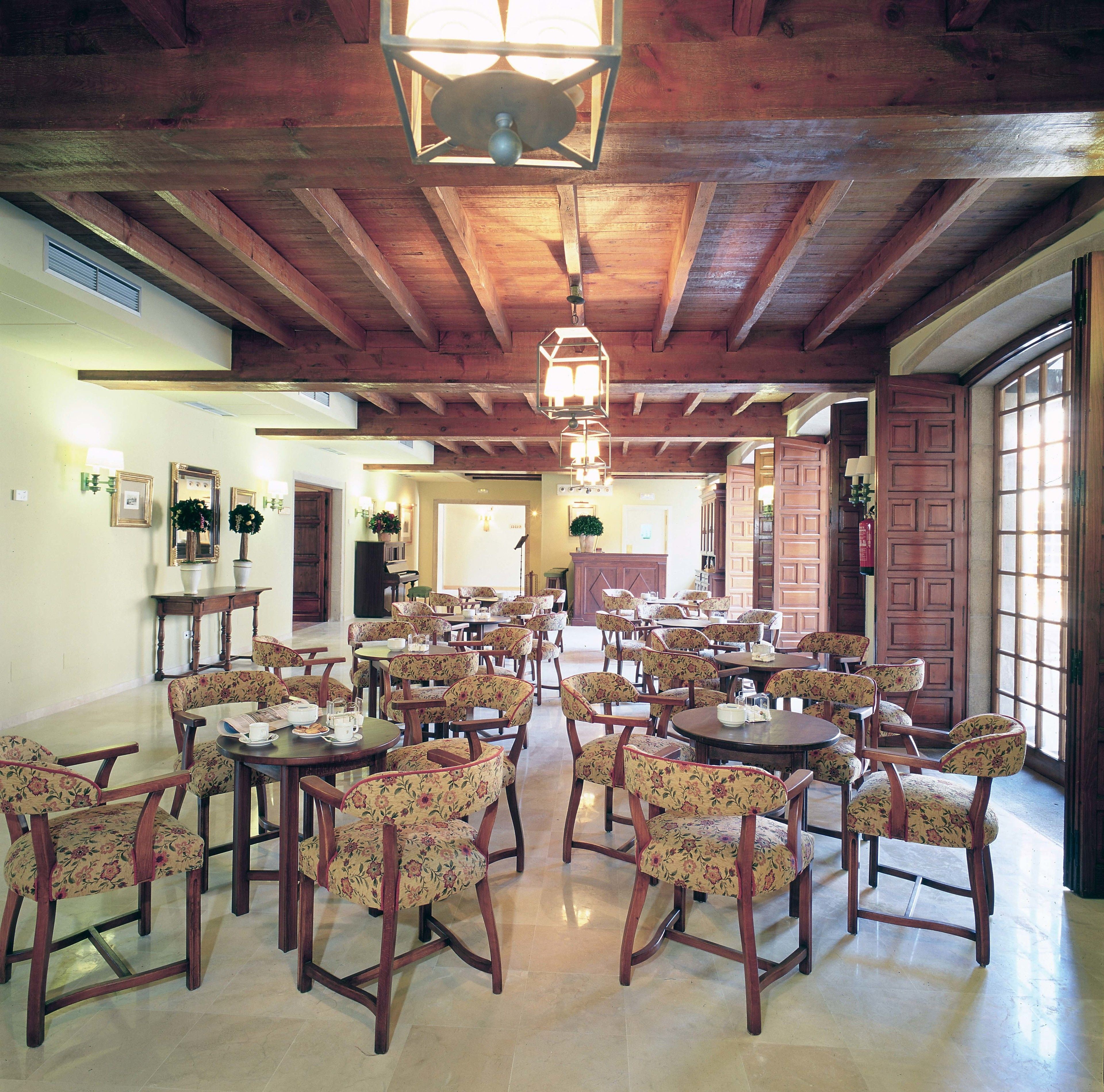 Un comedor amplio con mesas de madera y sillas tapizadas con motivos florales, que cuenta con vigas de madera a la vista y una gran lámpara de araña.