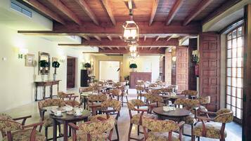 Un comedor amplio con mesas de madera y sillas tapizadas con motivos florales, que cuenta con vigas de madera a la vista y una gran lámpara de araña.