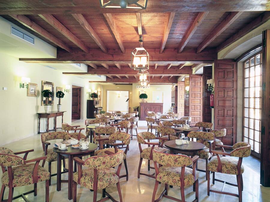 Un comedor amplio con mesas de madera y sillas tapizadas con motivos florales, que cuenta con vigas de madera a la vista y una gran lámpara de araña.